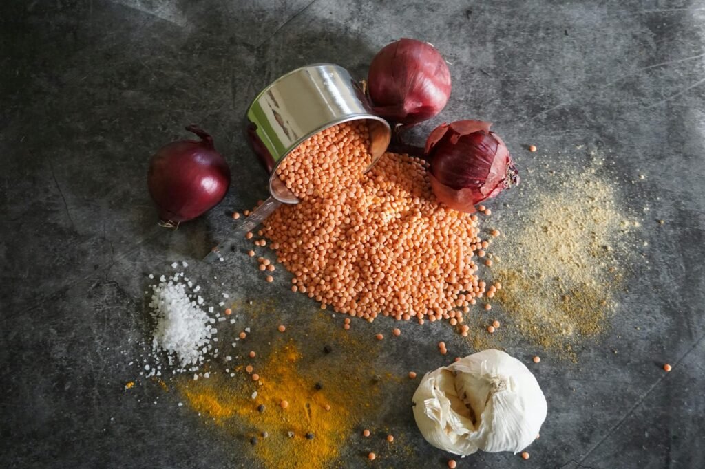 A spread of low-cost vegan pantry ingredients on a kitchen counter, including onions, potatoes, lentils, oats, canned beans, tomato paste, coconut milk, and pea